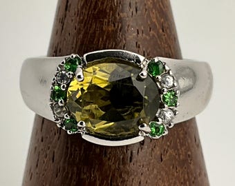 May include: A silver ring featuring a large, oval-cut, olive-green gemstone as the centerpiece. The ring is accented with smaller green and white gemstones around the main stone, creating a halo effect. The ring is displayed on a wooden stand.
