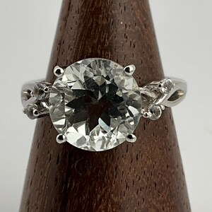 May include: A silver ring featuring a large, clear, round gemstone surrounded by smaller, clear gemstones. The ring is displayed on a wooden stand.
