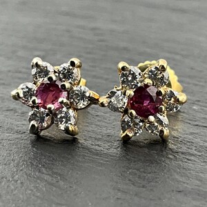 May include: A pair of gold stud earrings, each featuring a central round-cut deep pink gemstone surrounded by six small, clear, round-cut white stones arranged in a floral cluster pattern, set in yellow gold metal prongs.