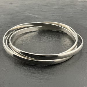 May include: Three silver bangles interlocked, creating a layered effect. The smooth, polished surface reflects light, highlighting the rounded edges. The bangles are displayed on a dark, textured surface.
