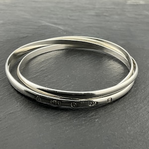 Può includere: Tre braccialetti d'argento intrecciati, su una superficie scura. I braccialetti sono lisci e lucenti, con piccoli segni su uno di essi. I gioielli sono semplici ed eleganti.