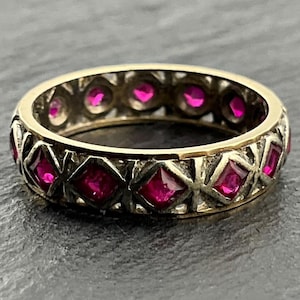 May include: A gold band ring featuring a continuous row of princess-cut, vibrant pinkish-red gemstones set within a textured, dark-toned metal framework that creates a repeating diamond-shaped pattern around the entire circumference.