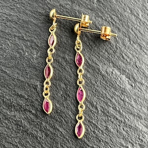May include: A pair of gold drop earrings featuring three marquise-cut red gemstones set in a vertical chain design. Each earring includes a post back closure with a small gold ball at the top, connecting to the dangling stones in a linear arrangement.