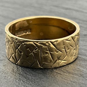 Könnte beinhalten: Ein breiter Goldring mit einer texturierten Außenfläche, die mit zahlreichen kleinen, sich überschneidenden linearen Einkerbungen und Markierungen versehen ist. Die Innenseite des Rings ist glatt und poliert und zeigt sichtbare Punzierungen.