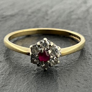 May include: A gold ring featuring a central round-cut red ruby surrounded by six small round-cut clear diamonds in a floral cluster setting. The band is smooth and polished gold, connecting to the central gemstone arrangement at the top of the ring.