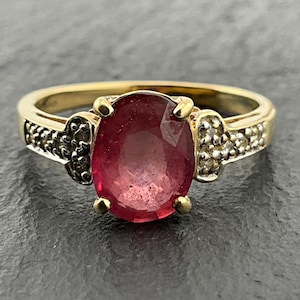 May include: A gold ring featuring a large, oval-cut, deep red gemstone set in the center. The band is accented on both sides by small, clear, sparkling stones embedded in the gold metal, creating a detailed and polished appearance.