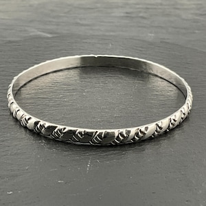May include: A silver bangle bracelet with a repeating heart pattern around the band. The bracelet is made of a shiny metal and has a simple, elegant design. The bracelet is shown on a dark surface.