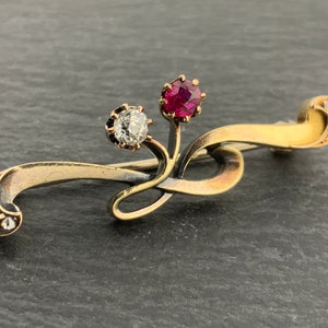 Könnte beinhalten: Eine goldene Brosche mit einem zarten, wirbelnden Design. Die Brosche hat einen roten Edelstein und einen klaren Edelstein.