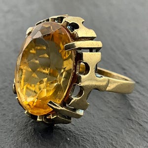 Könnte beinhalten: Ein goldener Ring mit einem großen, oval geschliffenen, facettierten bernsteinfarbenen Edelstein, der in einer dekorativen, durchbrochenen Metallfassung sitzt. Die Ringschiene ist glatt und verjüngt sich nach hinten, mit einer markanten, gekerbten Kante.
