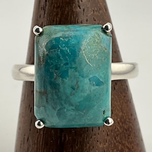 May include: A rectangular turquoise gemstone ring with a silver band. The turquoise stone is a vibrant blue-green colour and is set with four small silver accents. The ring is displayed on a wooden stand.