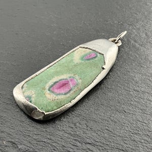May include: A rectangular pendant with a silver frame. The pendant features a green stone with pink and white patterns. The stone is set in a silver bezel with a textured edge and a small loop for a chain.