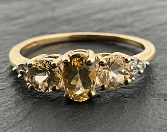May include: A gold ring featuring a central oval-cut yellow gemstone flanked by two round-cut yellow gemstones. The band is thin and polished gold, with small clear accent stones set into the shoulders on both sides of the main setting.