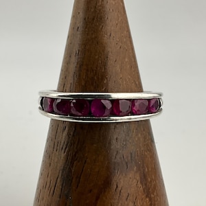 Może przedstawiać: Srebrny pierścionek z siedmioma okrągłymi, głęboko czerwonymi kamieniami szlachetnymi. Kamienie są ułożone w rzędzie w polerowanej srebrnej oprawie. Pierścionek jest prezentowany na drewnianym stojaku na neutralnym tle.