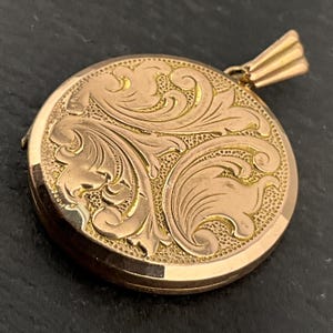 May include: A round, gold-colored locket with an ornate floral design. The locket features a detailed, engraved pattern of swirling leaves and vines. The bail is a simple, ribbed design. The locket is closed, and the overall appearance is vintage.