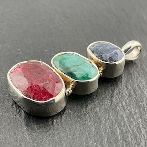 May include: A silver pendant with three oval gemstones: a deep red ruby, a vibrant green emerald, and a dark blue sapphire. The gemstones are set in individual silver bezels, connected in a row, with a silver loop for a chain.