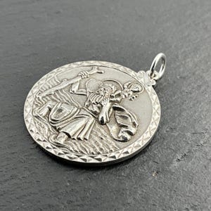 Könnte beinhalten: Ein silberner runder Anhänger mit einem detaillierten Relief von St. Christophorus, der ein Kind durch das Wasser trägt. Der Anhänger hat einen strukturierten Rand und eine kleine Schlaufe für eine Kette. Das Design ist aufwendig und das Metall hat eine polierte Oberfläche.