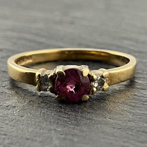 May include: A gold ring featuring a central round-cut deep pink gemstone flanked by two smaller clear round-cut stones. The band is smooth and polished, with the setting holding the three stones securely in place with small metal prongs.