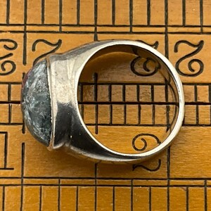 May include: A silver ring with a large, oval-shaped, green and black gemstone. The ring is set on a wooden ruler.