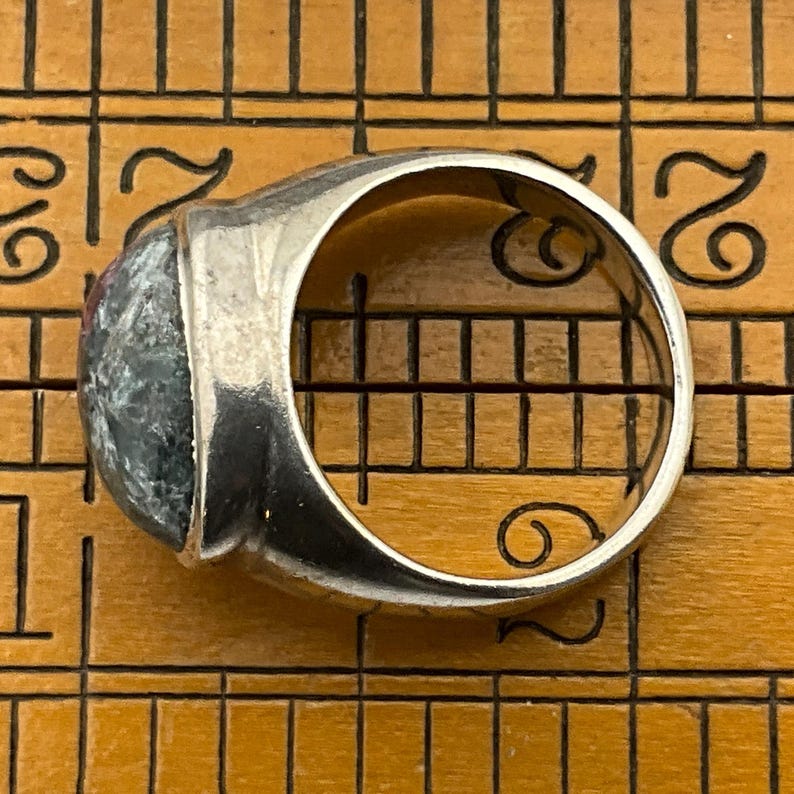 May include: A silver ring with a large, oval-shaped, green and black gemstone. The ring is set on a wooden ruler.