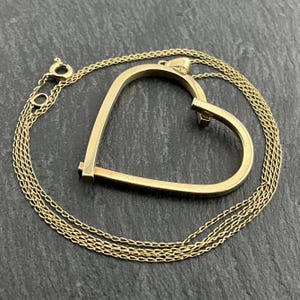 May include: A gold-colored necklace featuring a heart-shaped pendant. The pendant is a stylized open heart design, with a small gap at the top. The chain is a delicate, linked design. The necklace is set against a dark, textured background.