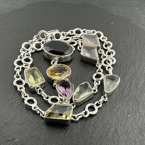 May include: A silver necklace with a variety of colourful gemstones in different shapes. The gemstones include black, yellow, purple, and green stones. The necklace has a chain-link design and is set against a dark background.