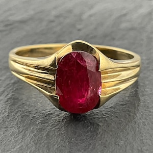 Puede incluir: Un anillo de oro con una piedra preciosa de color rojo intenso de corte ovalado en el centro. La banda está hecha de oro amarillo pulido y presenta una serie de tres ranuras horizontales a cada lado del engaste, hacia la piedra.