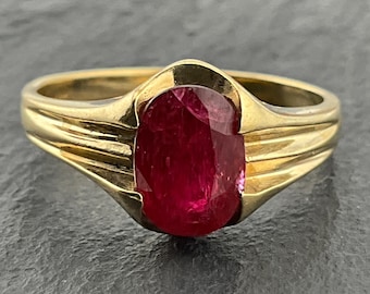 Puede incluir: Un anillo de oro con una piedra preciosa de color rojo intenso de corte ovalado en el centro. La banda está hecha de oro amarillo pulido y presenta una serie de tres ranuras horizontales a cada lado del engaste, hacia la piedra.