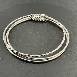 Könnte beinhalten: Drei silberne Armreifen, zwei glatt und einer mit gedrehtem Seilmuster, sind durch kleine silberne Ringe verbunden. Das Schmuckstück wird auf einer dunkelgrauen Oberfläche präsentiert. Ein klassisches Accessoire.