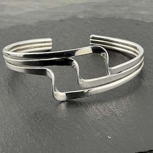 Puede incluir: Un brazalete de plata con un diseño ondulado abstracto. El brazalete está hecho de tres bandas de plata entrelazadas.