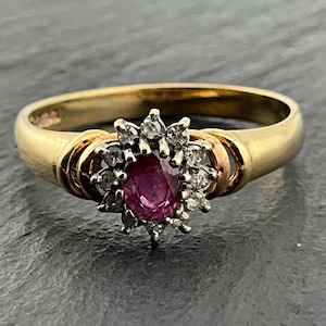 May include: A gold ring featuring a central oval-cut deep pinkish-red gemstone surrounded by a cluster of small, clear, round-cut stones. The band is smooth and polished, with a setting that elevates the central stone arrangement.