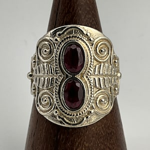 May include: A silver ring with two oval-shaped, dark red gemstones set in a detailed, ornate design.