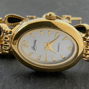 Peut inclure: Une montre-bracelet ovale dorée avec un cadran blanc, des chiffres romains noirs et des accents de pierres précieuses noires sur le bracelet. Le cadran de la montre indique "Gemsyk Quartz".