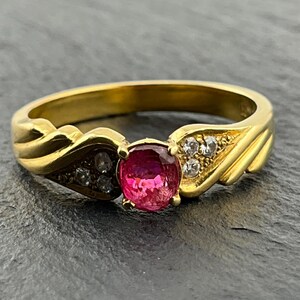 Op de afbeelding: Een gouden ring met een centrale ovaal geslepen, levendig roze edelsteen in een zetting met vier pootjes. De ringband is aan beide zijden versierd met een driehoekig gedeelte met drie kleine, heldere, rond geslepen stenen in het goud.