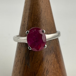 May include: A silver ring with a large, oval-cut ruby gemstone. The ruby is a deep red colour and is held by four prongs. The ring's band is polished silver. The ring is displayed on a wooden stand.