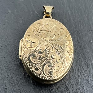 May include: An oval-shaped gold locket pendant featuring an intricate engraved floral and scrollwork pattern across the front surface. The piece includes a small, triangular-shaped gold bail at the top for attachment to a chain or necklace.