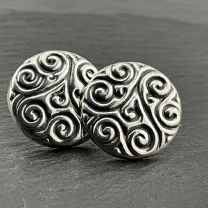 May include: Two silver-colored, round stud earrings with an ornate, swirling pattern. The earrings are highly detailed, with a raised design against a darkened background. The earrings are displayed on a dark surface.