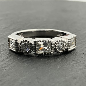 Op de afbeelding: Een zilveren bandring met vijf heldere zirkonia-stenen. De stenen zijn gezet in een bezel-setting en worden omringd door een decoratieve rand. De ring is een klassiek en elegant ontwerp.