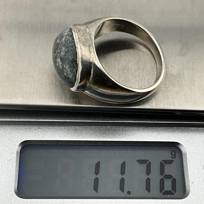 May include: A silver ring with a dark green and black gemstone. The ring is on a digital scale that reads 11.76 grams.