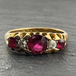 May include: A gold ring featuring a central oval-cut red ruby flanked by two smaller round-cut diamonds and two outer oval-cut red rubies. The stones are set in a yellow gold band with a scalloped metal design surrounding each individual gemstone.