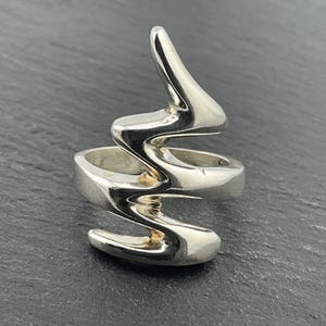 Puede incluir: Un anillo de plata con un diseño único y abstracto que se asemeja a un rayo. El anillo está hecho de metal liso y pulido y tiene una estética moderna y minimalista.