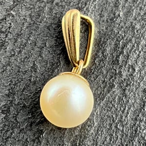 May include: A gold pendant featuring a single, round, cream-coloured pearl suspended from a polished gold bail. The bail has a vertical, ribbed design that connects to the pearl setting. The pearl has a smooth, lustrous surface and a spherical shape.