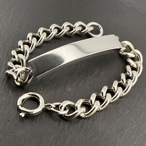 Könnte beinhalten: Ein silberfarbenes Panzerkettenarmband mit einer rechteckigen Platte in der Mitte. Das Armband hat einen Karabinerverschluss. Das Armband wird auf einer dunkelgrauen Oberfläche gezeigt.