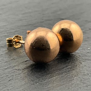 May include: A pair of polished rose gold ball stud earrings. Each earring features a smooth, spherical design with a reflective metallic surface. The earrings include a standard post and a secure butterfly back closure, showcasing a simple, rounded shape.