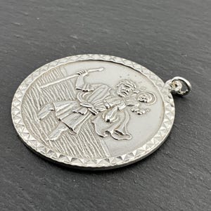 Puede incluir: Pendiente de plata con un relieve detallado de San Cristóbal llevando a un niño. El pendiente tiene un borde texturizado y un bucle para sujetar una cadena.