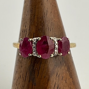 May include: A gold ring featuring three oval-cut, deep red gemstones, likely rubies, set in a row. Small, clear stones accent the spaces between the rubies. The ring is displayed on a wooden stand.