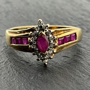 May include: A gold ring featuring a central marquise-cut ruby surrounded by a cluster of small round diamonds. The band includes channel-set square-cut rubies on both sides, extending from the central setting along the gold shank of the ring.