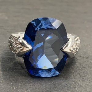 Puede incluir: Un anillo de plata con una gran piedra preciosa azul oscuro engastada en forma de cojín. El anillo tiene una banda delicada con detalles de diamantes.