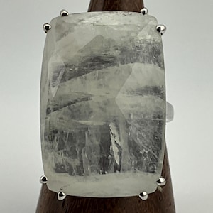 Puede incluir: Un anillo grande y rectangular con una piedra preciosa pálida y translúcida. La piedra está facetada y engastada con pequeños detalles plateados en cada esquina. El anillo se exhibe sobre un soporte de madera sobre un fondo blanco.
