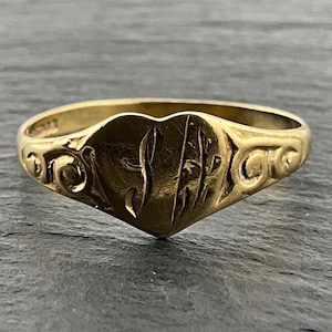 May include: A gold ring featuring a heart-shaped face with engraved initials that appear to be SH. The band is decorated with ornate scrollwork patterns on both sides, extending from the central heart element to the back of the ring.