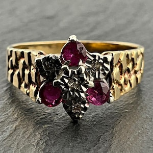 Puede incluir: Un anillo de oro con una banda texturizada estilo pepita y un adorno central en forma de flor. El adorno está engastado con tres rubíes redondos de color rosa intenso y pequeñas piedras claras en un engaste de metal blanco.